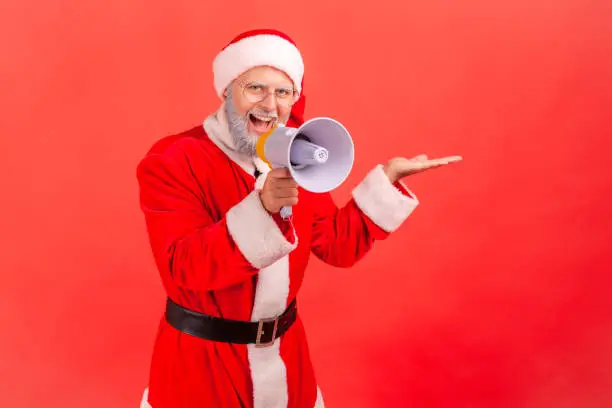 Smiling happy elderly man in santa claus costume showing freespace for advertisement and talking into loudspeaker, holidays promotion. Indoor studio shot isolated on red background