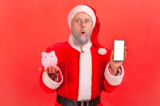 Photo of Shocked surprised santa claus holding smartphone with empty screen and piggy bank, cashback, online wallet.