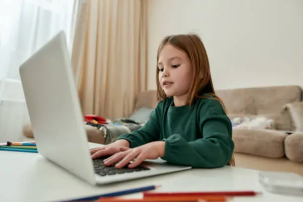 A cute small girl enjoys chatting with her friends via a laptop while sitting alone in a huge bright guestroom with her pencils all around a table alone during distance education. Home schooling