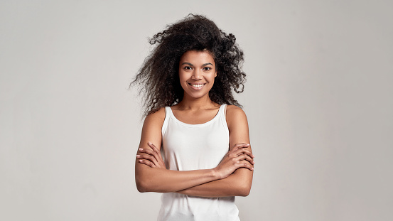 Portrait of young african american woman with curly hair wearing white shirt smiling at camera while standing with arms crossed isolated over grey background. Front view. Web Banner