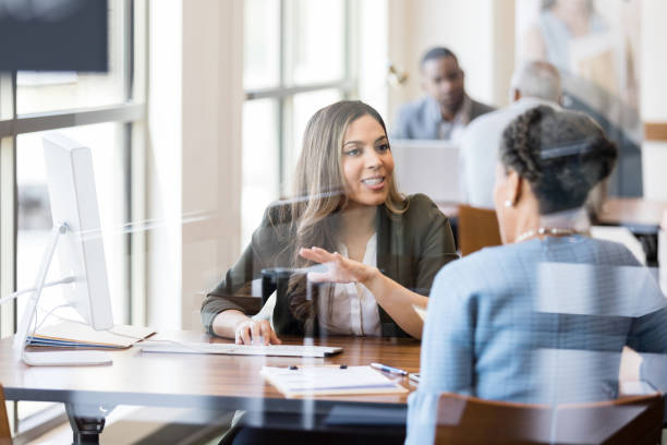 Banker discusses banking services to new customer A mid adult female bank employee gestures as she explains banking services to a female customer. banking stock pictures, royalty-free photos & images