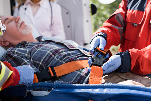 Selective focus of paramedic locking belts of stretcher near elderly patient and colleagues outdoors