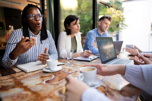 Diverse group of business people sitting together, having a business meeting at the restaurant. Businessman and businesswoman using laptop. Young African ethnicity businesswoman explaining.