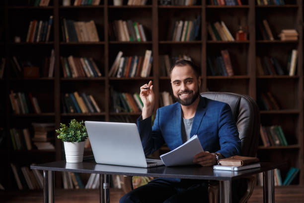 молодой бородатый бизнесмен улыбается и смотрит в кадр среди книг в библиотеке за столом перед ноутбуком - general manager men business person стоковые фото и изображения