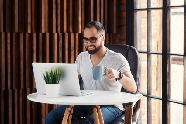 istock Young successful bearded businessman freelancer in office coworking. Happy man programmer developer in a white t shirt smiling and drinking coffee at the Desk with a laptop 1281367208