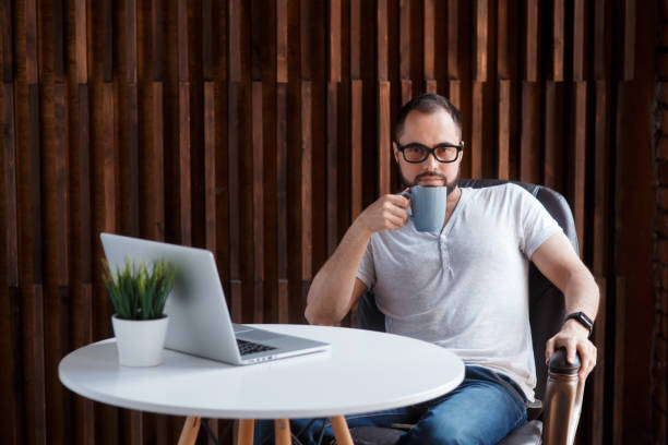 istock Young successful bearded businessman freelancer in office coworking. Happy man programmer developer in a white t shirt smiling and drinking coffee at the Desk with a laptop 1281367212