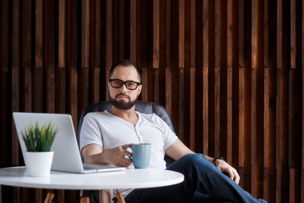 istock Young successful bearded businessman freelancer in office coworking. Happy man programmer developer in a white t shirt smiling and drinking coffee at the Desk with a laptop 1281367216
