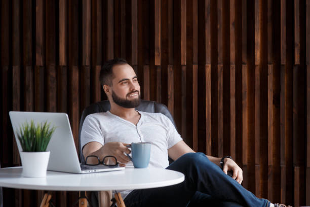 istock Young successful bearded businessman freelancer in office coworking. Happy man programmer developer in a white t shirt smiling and drinking coffee at the Desk with a laptop 1281367220