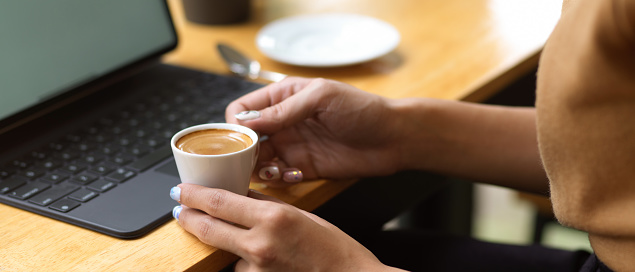 Side view of female hands holding coffee cup while sitting at workplace with digital tablet