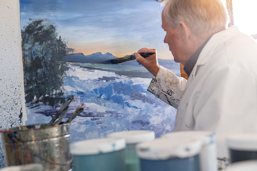 Senior man painting impressionistic art in his studio while sitting.