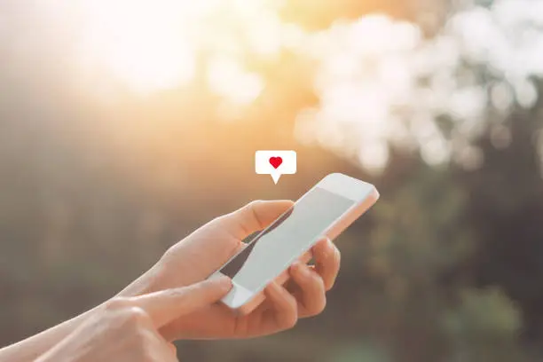 Photo of Close up woman hand hold using smart phone with heart icon at outdoor park street background.