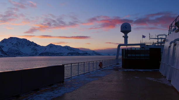 Sørøysundet, Norway - 03/02/2019: Upper deck of Hurtigruten cruise ship MS Trollfjord with orange colored clouds in the evening light and snow-covered rugged mountains.