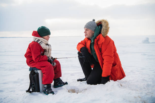 A little boy in a red jumpsuit and scarf is fishing in an ice hole with his father stock photo