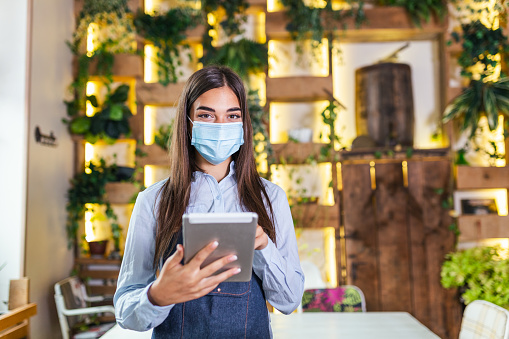 Portrait of beautiful waitress wearing protective face mask while holding touchpad and looking at the camera in a pub or restaurant during COVID-19 epidemic.