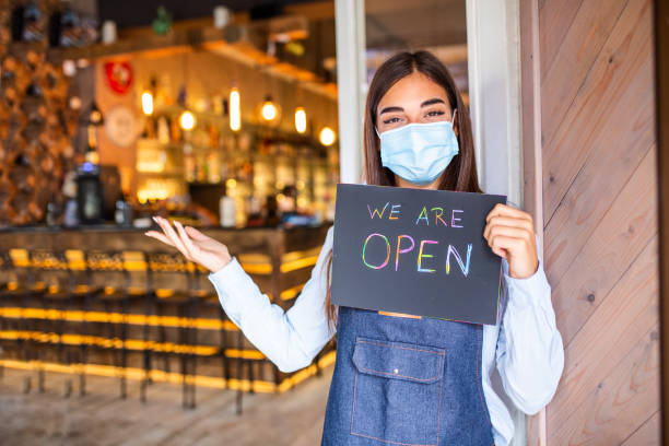 istock Happy female waitress with protective face mask holding open sign while standing at cafe or restaurant doorway, open again after lock down due to outbreak of coronavirus covid-19 1281392664