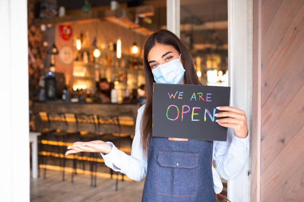 istock Happy female waitress with protective face mask holding open sign while standing at cafe or restaurant doorway, open again after lock down due to outbreak of coronavirus covid-19 1281392709