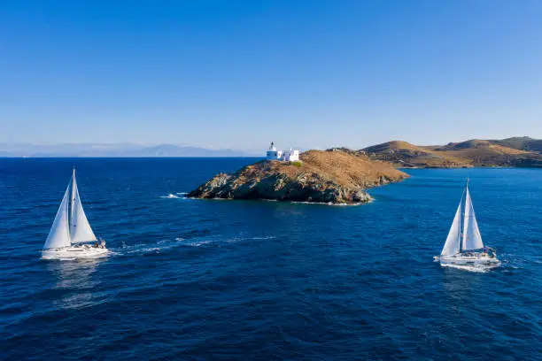 Sailing. Sailboats with white sails, rippled sea background, Lighthouse on a cape. Greece, Kea Tzia island. Summer holidays in Aegean sea. Aerial drone view
