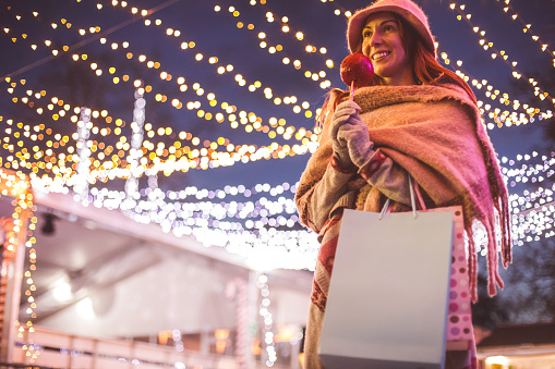 Low angle view of happy woman in winter clothes holding shopping bags and walking in the street with Christmas lights on the background.