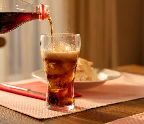 Photo of Pouring Cola from Bottle into Glass and Fizz with Ice Cubes on Table Against Blurred Livinroom  Background