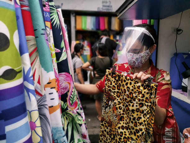 Divisoria, Manila, Philippines - A woman wearing a face mask and shield shops for bedsheets at a local stall at a street market. Divisoria, Manila, Philippines - Oct 2020: A woman wearing a face mask and shield shops for bedsheets at a local stall at a street market. divisoria market stock pictures, royalty-free photos & images