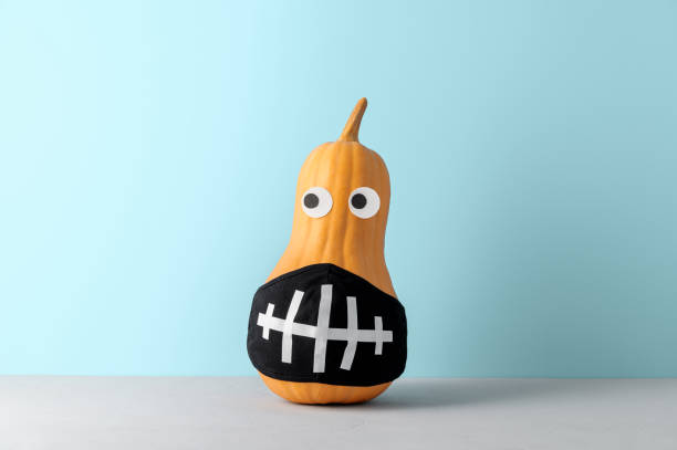Funny pumpkin with face protective mask on pastel blue background with copy space.