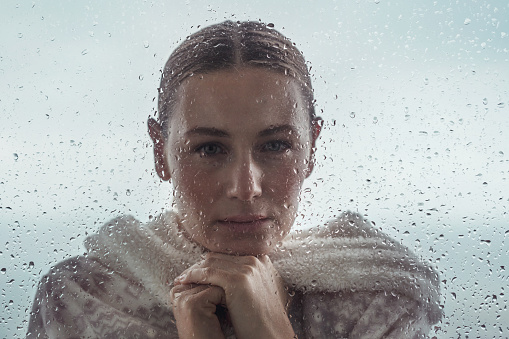 Portrait of a sad woman looking from outdoors into the house through a window covered with raindrops, authentic portrait of a calm girl