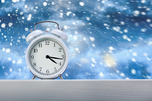 Alarm clock on wooden windowsill in snowy weather on winter background. Winter time concept.
