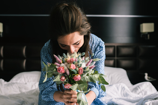 Young girl rejoices at the presented bouquet of flowers. A woman in a blue dress sits on the bed and sniffs roses. The concept of wedding, flower delivery and love.