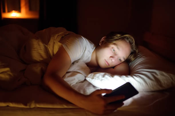 nastolatek chłopiec rozmawia przez telefon, pisanie na blogu lub czytanie sieci społecznych zamiast spać. młody facet leży w łóżku i ogląda klipy przez smartfona. nadużywanie i uzależnienie dzieci od gadżetów. - teenage boys zdjęcia i obrazy z banku zdjęć