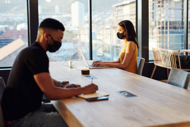 A bit of distance benefits everyone Shot of a masked young businesswoman and businessman working apart from each other at a table in a modern office adaptation concept stock pictures, royalty-free photos & images