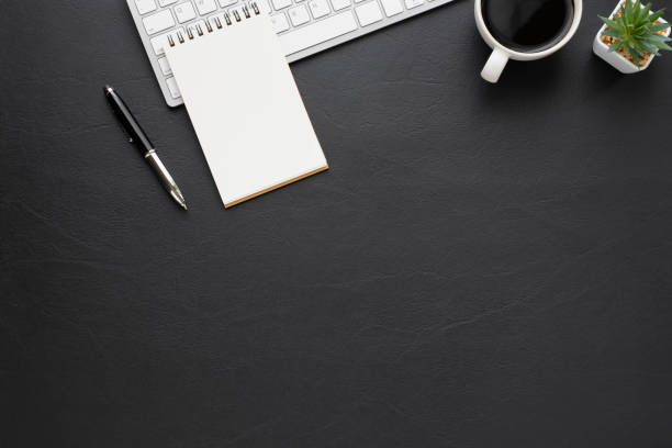 istock Top view above of Black leather office desk table with keyboard, notebook and coffee cup with equipment other office supplies. Business and finance concept. Workplace, Flat lay with blank copy space. 1281464775