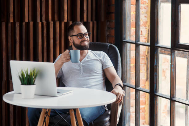 istock Young successful bearded businessman freelancer in office coworking. Happy man programmer developer in a white t shirt smiling and drinking coffee at the Desk with a laptop 1281465950