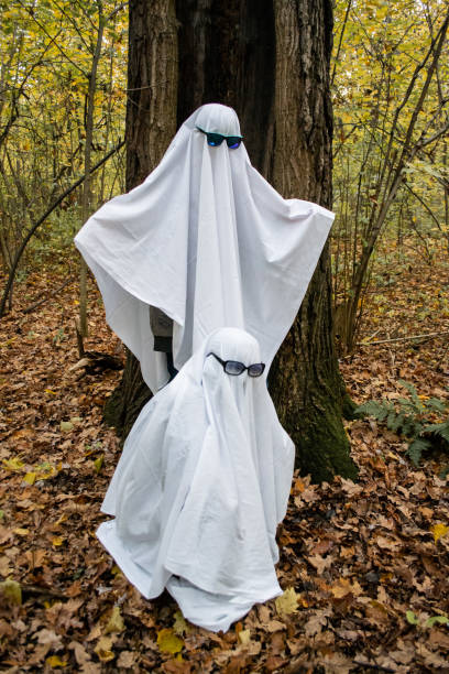 istock Ghost challenge in the forest or park. Two unrecognizable people teenagers disguised as ghosts with white bed sheets and sunglasses. One sits, another stands, near big old tree 1281469047