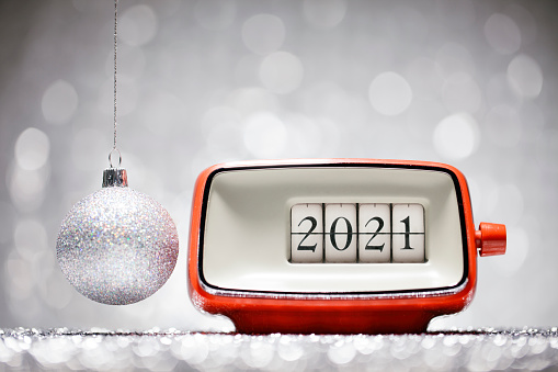 Retro alarm clock showing the number 2021. A Christmas ornament hanging besides it. White defocused lights in the background.