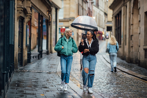 Two female students walking through the city centre of Newcastle. It is raining and they are holding an umbrella.