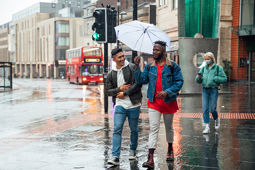 A young man carrying an umbrella walks through the city centre with his friend to keep them as dry as possible. There is a young woman in the background wearing a protective mask.