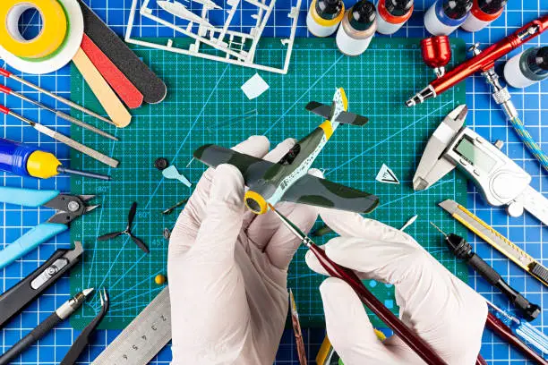 desktop view from above of assembly and painting of retro scale model fighter plane concept background. modeling tools airbrush gun paint kit parts blue green cutting mat knife and brush on work desk