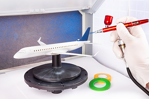 paint job with red metal airbrush spray paint gun on red scale model on modern white blue passenger airplane plane in paint booth. hobby industry concept background