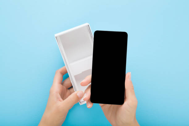 istock Young woman hand taking out new smartphone from white box on light blue table background. Pastel color. Closeup. Empty place for text on black screen. Point of view shot. Top down view. 1281488193