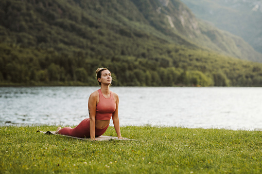 A woman is doing yoga outside, near the scenic lake. The Cobra pose.  She is wearing a yoga sportswear and practicing on a yoga mat. Copy space. Bhujangasana