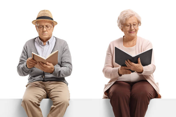 istock Elderly man and woman sitting on a white panel reading books 1281489800