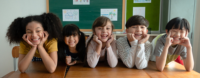 Portrait of happy elementary international students sitting on desk touches cheeks with both hands and looking at camera in classroom.Web banner panorama with adorable school kids.Education concept.