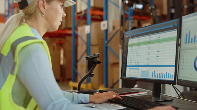 Professional Female Worker Wearing Hard Hat Uses Computer with Inventory Status Checking and Delivery Software in the Retail Warehouse full of Shelves with Goods. Delivery, Distribution Center