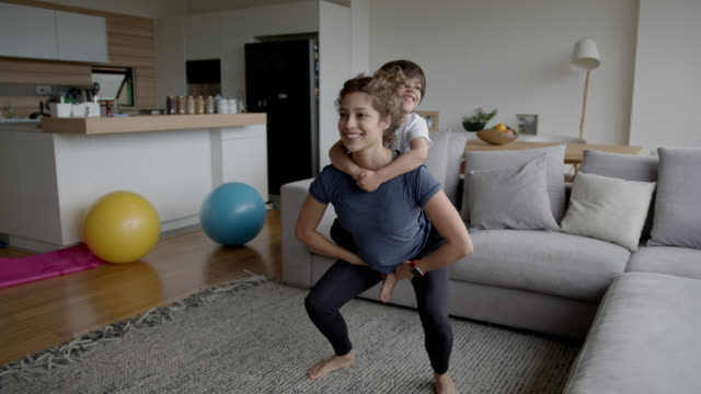 Young mother exercising at home carrying her son while doing squats both smiling - Fitness concepts