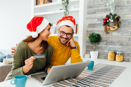 Young Caucasian couple having fun at home, celebrating winter festivities together, smiling and using a credit card to do some Christmas shopping online using a laptop