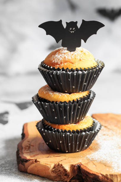 Halloween pumpkin muffins in black capsules decorated with cardboard bats. Festive Halloween cupcakes. Close-up view of delicious spooky halloween muffins on the grey background. Halloween recipes muffins de calabaza de halloween en cápsulas negras decoradas con murciélagos de cartón. cupcakes festivos de halloween. vista de primer plano de deliciosos y espeluznantes magdalenas de halloween sobre el fondo gris. recetas de halloween
