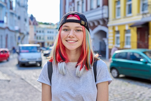 Portrait of fashionable hipster teenage girl with colored dyed hair in black cap and headphones, city street background. Lifestyle, fashion, trends, youth concept