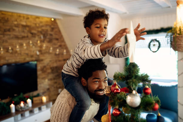 Small black boy decorating Christmas tree with his father and putting star on top. Happy African American father carrying his son on shoulders and helping him to put star on top of Christmas tree. traditional christmas stock pictures, royalty-free photos & images