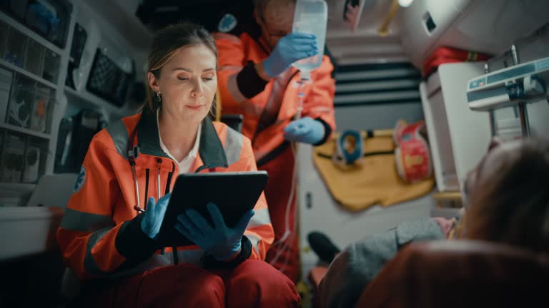 Female EMS Professional Paramedic Using Tablet Computer to Fill a Questionnaire for the Injured Patient on the Way to Hospital. Emergency Care Assistant Comforting the Patient in an Ambulance.