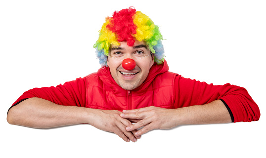 Smiling funny clown with a multi colored wig leaning over a billboard banner. Isolated on white. You can add extra white space with your message to the bottom.